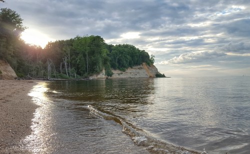 A serene beach scene with gentle waves, lush green cliffs, and a cloudy sky reflecting the sunlight.