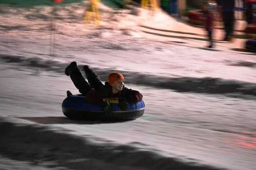 A child joyfully sledding down a snowy hill on a blue inner tube at night, wearing a helmet and gloves.