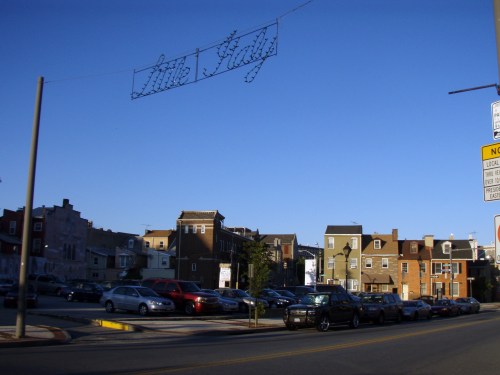 A street view of a neighborhood with a "Little Italy" sign overhead and parked cars along the road under a clear blue sky.