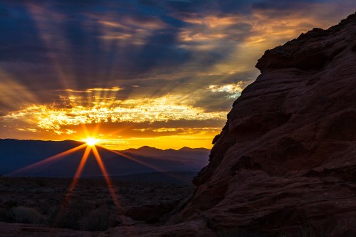 Sunset over rugged mountains, with rays of light shining through clouds and illuminating the rocky landscape.