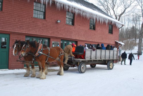 A horse-drawn wagon filled with people stands in front of a red barn, surrounded by snow and icicles.