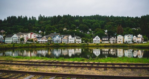 Colorful houses line a riverbank, reflecting in the calm water, with lush green hills in the background under a cloudy sky.
