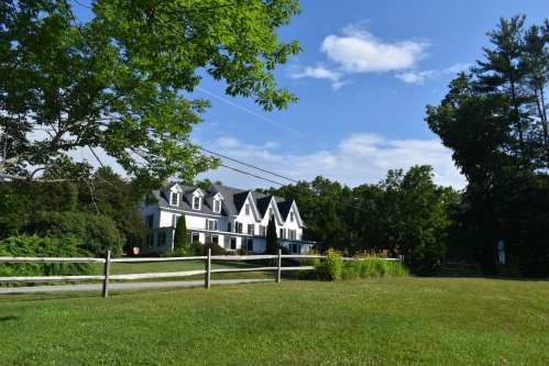 A white house with multiple gables surrounded by greenery and a wooden fence under a blue sky with clouds.