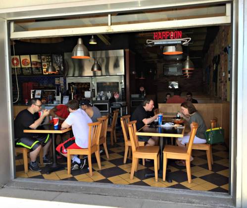 A casual restaurant scene with diners at tables, enjoying meals and drinks, with a visible kitchen in the background.