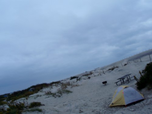 A tent set up on a sandy beach under a cloudy sky, with a picnic table in the background.