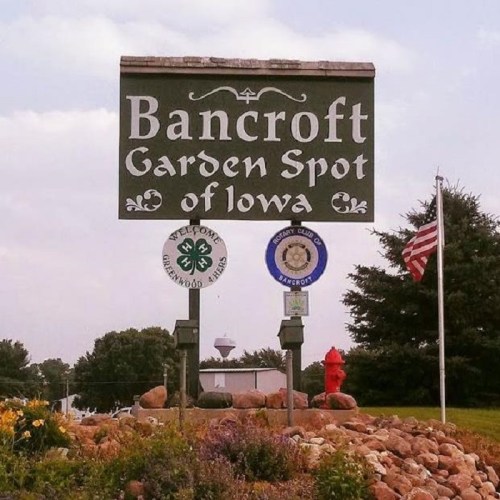 Sign for Bancroft, Iowa, reading "Bancroft Garden Spot of Iowa," with local symbols and an American flag nearby.