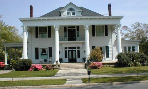 A large, white two-story house with columns, green shutters, and a manicured lawn, featuring colorful flowers in front.