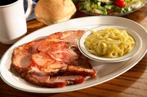 A plate of sliced ham with a side of fettuccine and a roll, accompanied by a salad in the background.