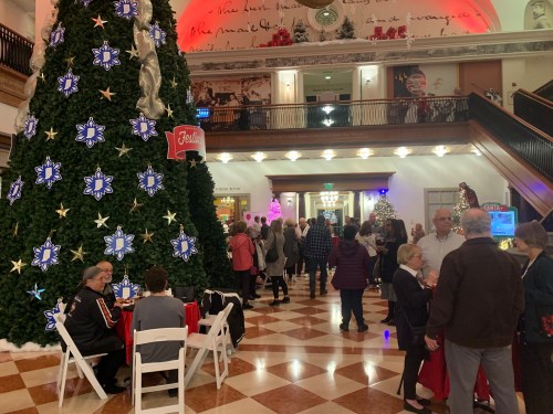 A festive indoor scene with a large Christmas tree, people mingling, and holiday decorations in a busy atrium.