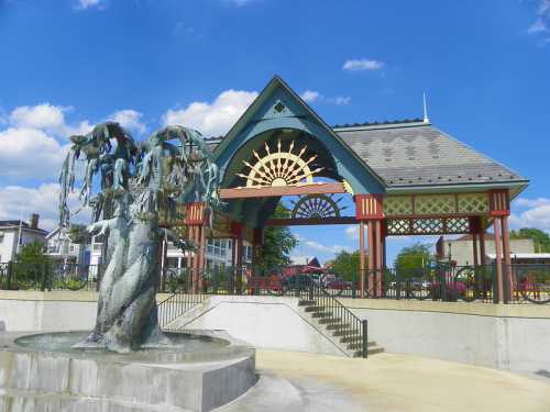 A decorative pavilion with a fountain featuring a sculpted tree, set against a blue sky with fluffy clouds.