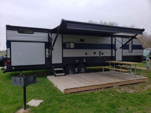 A parked travel trailer with an awning, next to a wooden deck and a grill, set in a grassy area.
