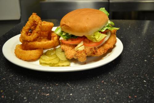 A plate with a crispy chicken sandwich topped with lettuce, tomato, and pickles, alongside onion rings.