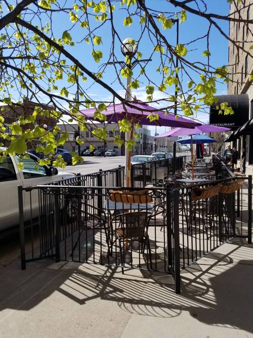 Outdoor café with tables and umbrellas, surrounded by greenery and a sunny street view.