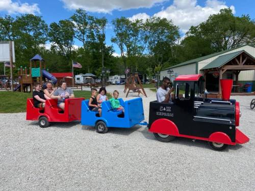 A small train ride with children and adults in colorful cars at a playground, surrounded by trees and a building.