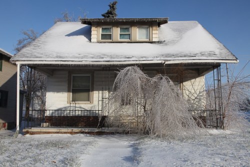 A snow-covered house with a sloped roof and a drooping tree in front, surrounded by a winter landscape.