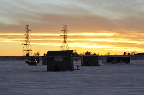 A snowy landscape at sunset, featuring power lines and small structures silhouetted against the colorful sky.