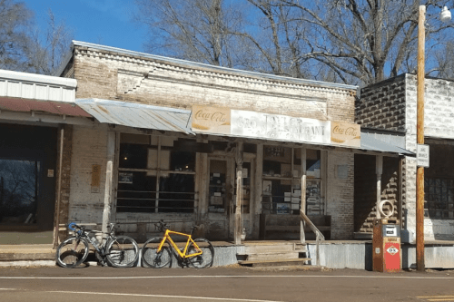 A vintage storefront with a Coca-Cola sign, featuring bicycles parked outside and a rustic wooden porch.