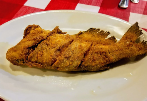 A whole fried fish on a white plate, served on a red and white checkered tablecloth.