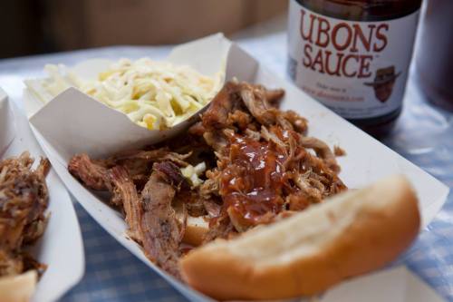 A plate of pulled pork with barbecue sauce, coleslaw, and a bun, alongside a bottle of Ubon's sauce.
