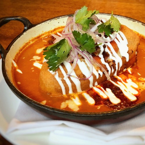 A bowl of rich, spicy sauce with a stuffed tortilla, topped with onions, cilantro, and a drizzle of cream.