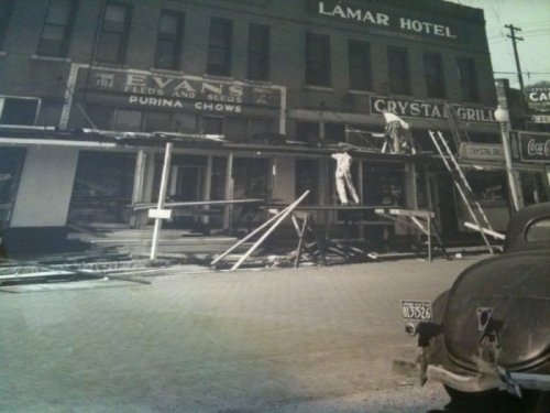 Historic black-and-white photo of a damaged building with scaffolding, featuring the Lamar Hotel and nearby shops.
