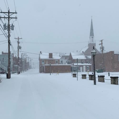 A snowy street scene with a church steeple in the background, power lines, and buildings partially obscured by snow.