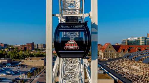 A close-up of a gondola on a large Ferris wheel against a clear blue sky and city skyline.