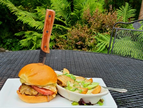 A burger with a knife in it, served alongside a dish of roasted potatoes and a fresh salad on a table outdoors.