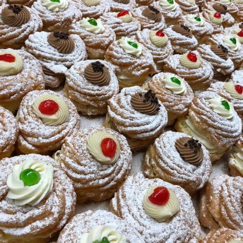 A close-up of a variety of cream-filled pastries topped with whipped cream and cherries, dusted with powdered sugar.