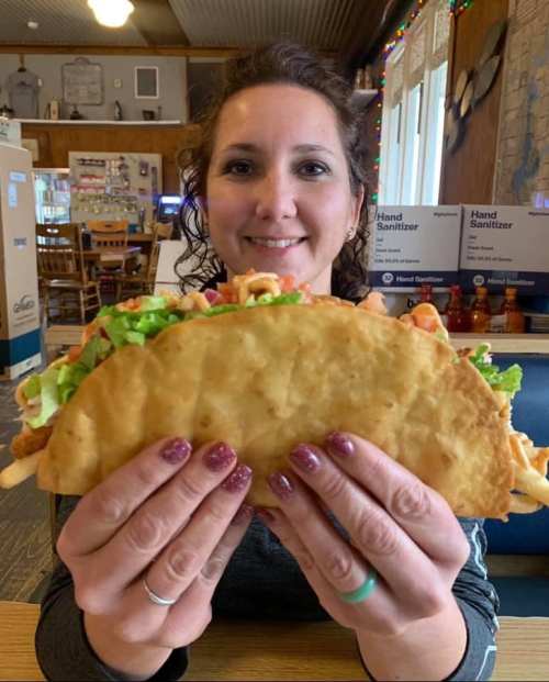 A woman holds a large taco filled with lettuce, tomatoes, and other toppings, smiling at the camera.