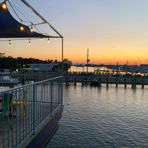 A serene waterfront view at sunset, with boats docked and lights glowing on a nearby deck.