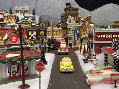 A festive miniature town scene with colorful buildings, cars, and holiday decorations, set in a snowy landscape.