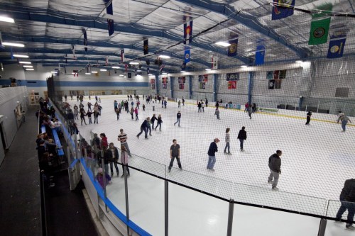 A busy indoor ice skating rink with people skating and socializing, surrounded by banners and a netted area.