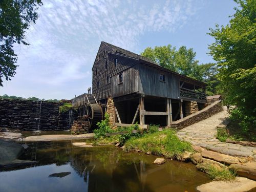 Historic wooden mill with a waterwheel, surrounded by greenery and a small pond under a blue sky.
