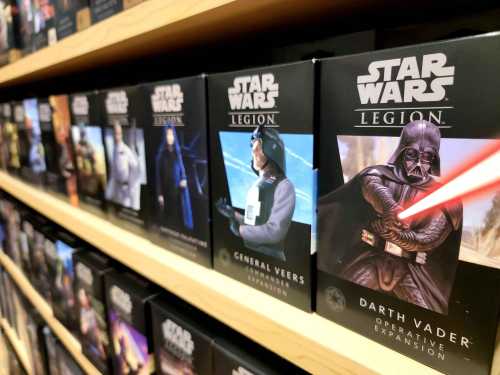 A shelf displaying various "Star Wars: Legion" game expansion boxes, featuring characters like Darth Vader and General Veers.