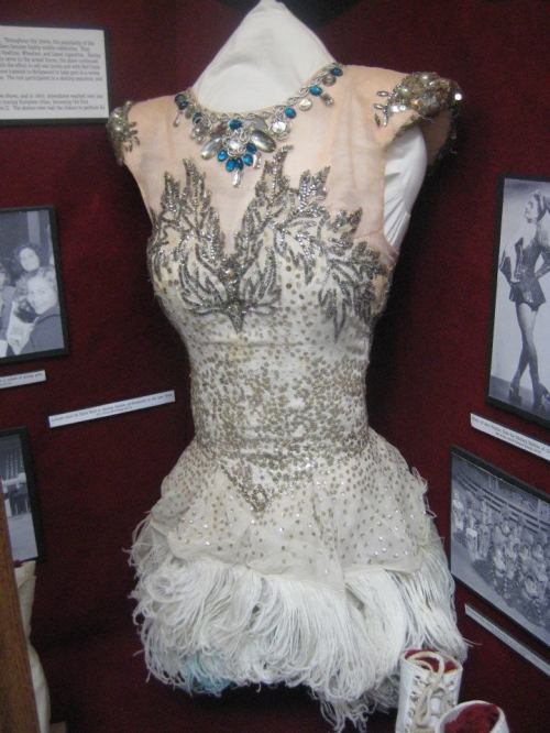 A glamorous dress adorned with beads and sequins, featuring a feathered hem, displayed in a museum setting.