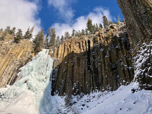 A frozen waterfall cascades down rocky cliffs, surrounded by snow-covered trees and a blue sky.