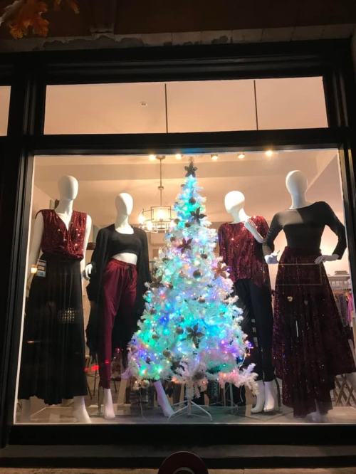 A store window display featuring mannequins in festive outfits around a decorated Christmas tree.