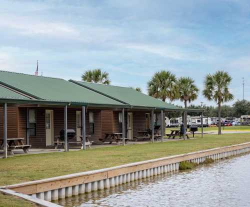 Row of cabins with green roofs beside a calm waterway, surrounded by palm trees under a cloudy sky.