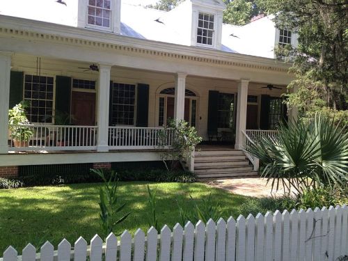 A charming white house with a porch, green shutters, and a white picket fence, surrounded by lush greenery.