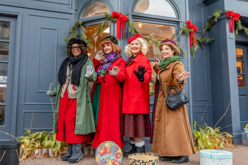 Four women in festive winter outfits pose cheerfully outside a decorated storefront, surrounded by holiday greenery.