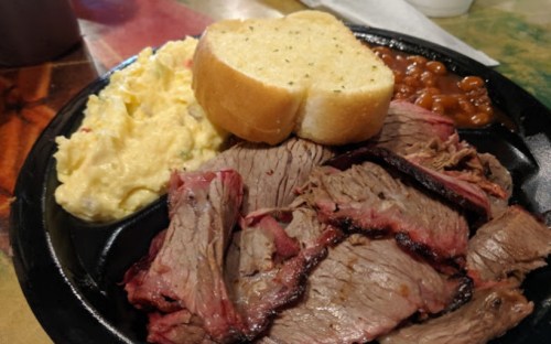 A plate of sliced brisket, creamy mashed potatoes, baked beans, and a slice of garlic bread.