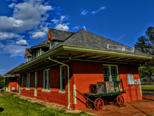 Historic red brick train station with green trim, surrounded by grass and under a blue sky with clouds.