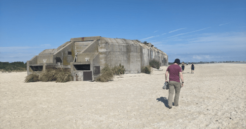 A person walks towards a weathered concrete bunker on a sandy beach, with another figure in the background.