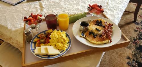 A breakfast tray with pancakes, bacon, scrambled eggs, cheese, fruit, and drinks, set on a table with a floral tablecloth.