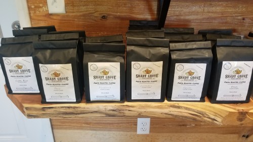 A display of black coffee bags labeled "Shady Grove" on a wooden shelf, showcasing various coffee blends.