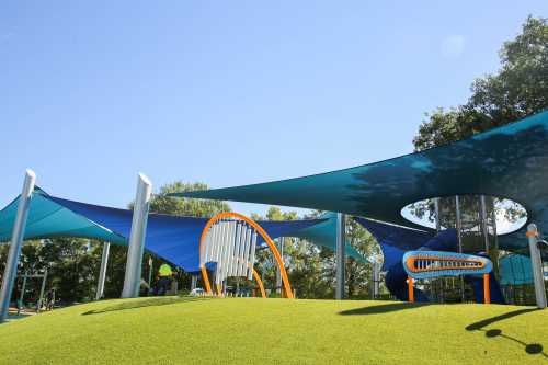 Colorful playground with large shade sails, grassy hill, and various play structures under a clear blue sky.