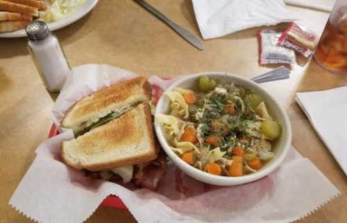 A sandwich with toasted bread and a bowl of chicken noodle soup with vegetables on a table.