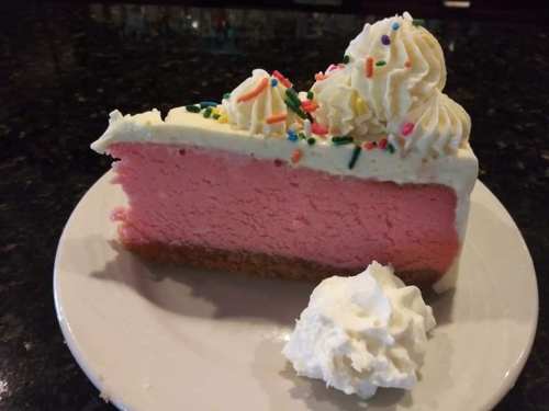 A slice of pink cheesecake topped with whipped cream and colorful sprinkles, served on a white plate with a dollop of whipped cream.