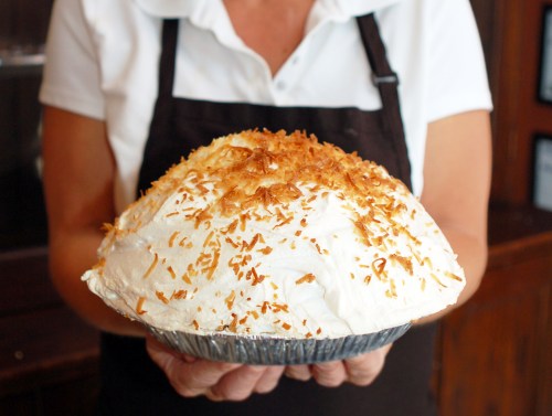 A person holds a large coconut cream pie topped with toasted coconut flakes.